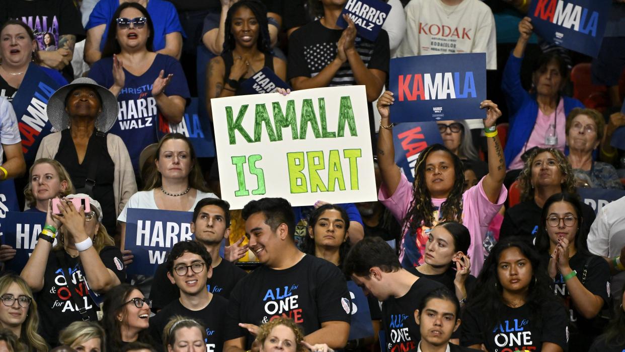 Supporters cheer for US presidential candidate Harris at a rally; a "Kamala is brat" sign stands out in th crowd