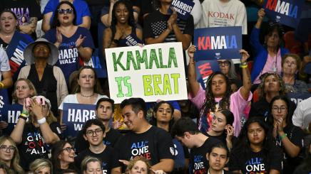 Supporters cheer for US presidential candidate Harris at a rally; a "Kamala is brat" sign stands out in th crowd